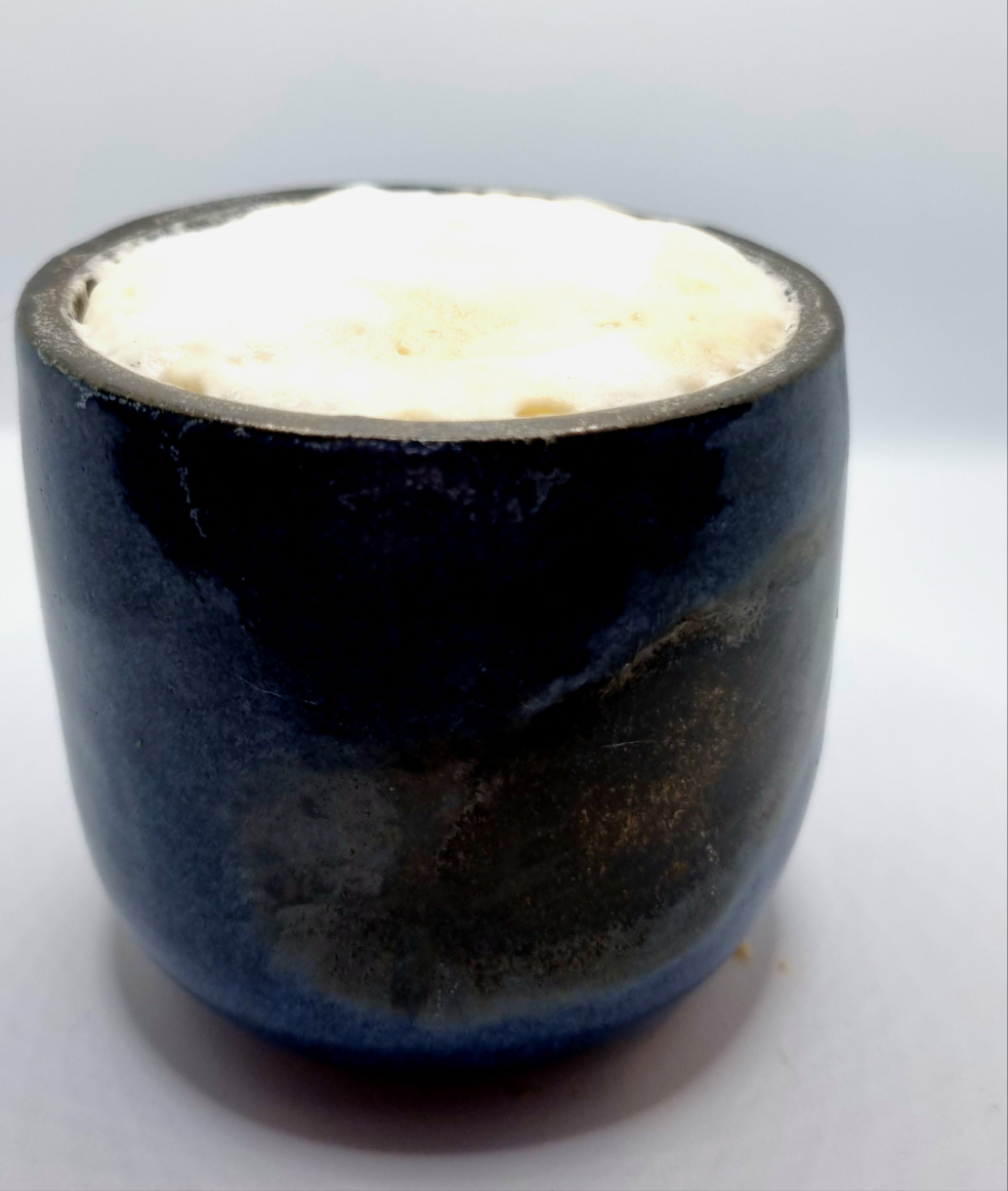 A dark blue & bronze ceramic tumbler with Cappuccino foam on a neutral background