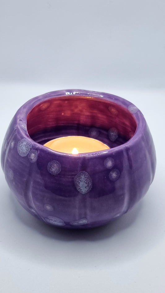 A purple, pumpkin-shaped tealight holder with a lit tealight inside on a neutral background
