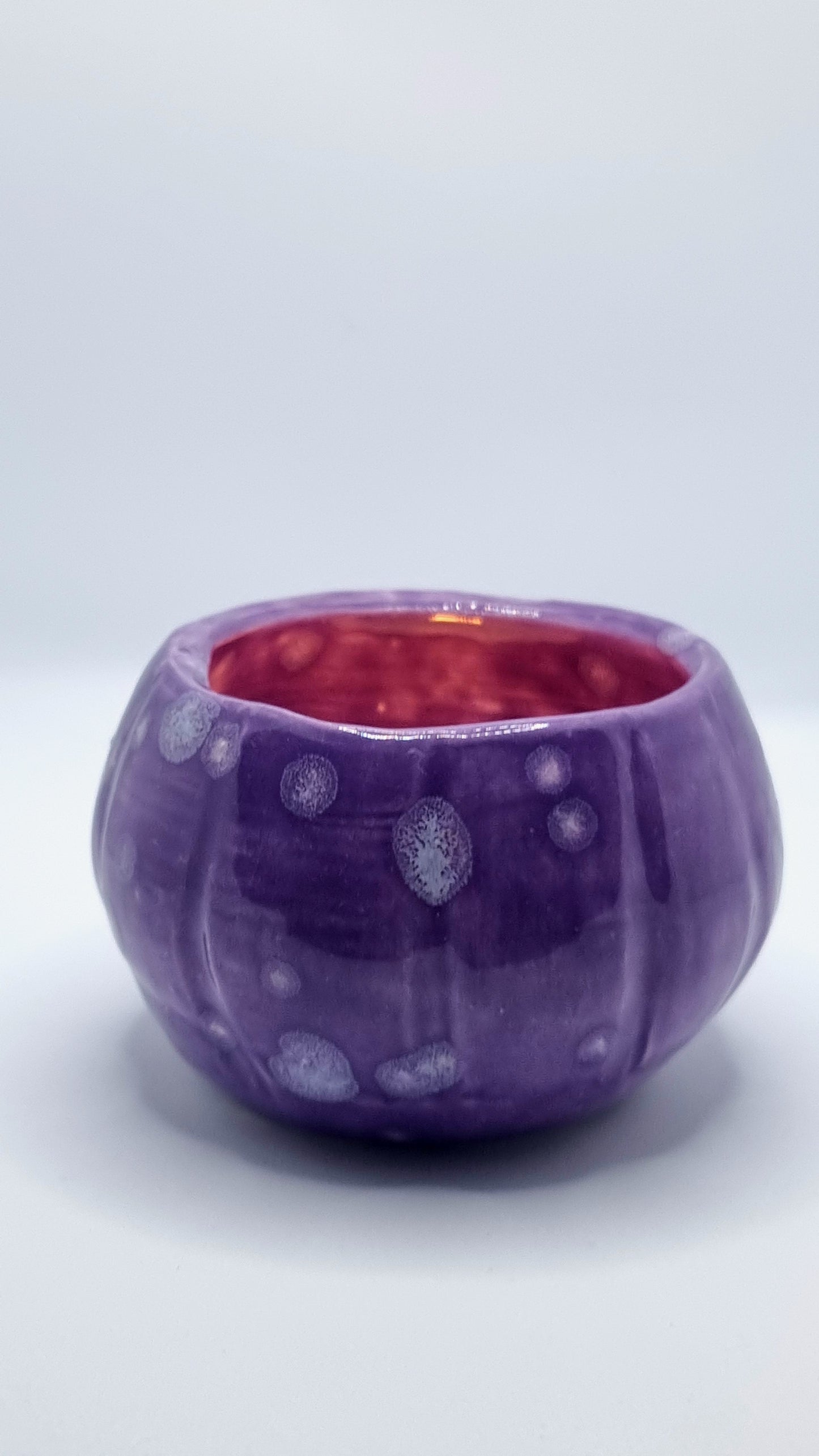 A purple, pumpkin-shaped tealight holder with a lit tealight inside on a neutral background
