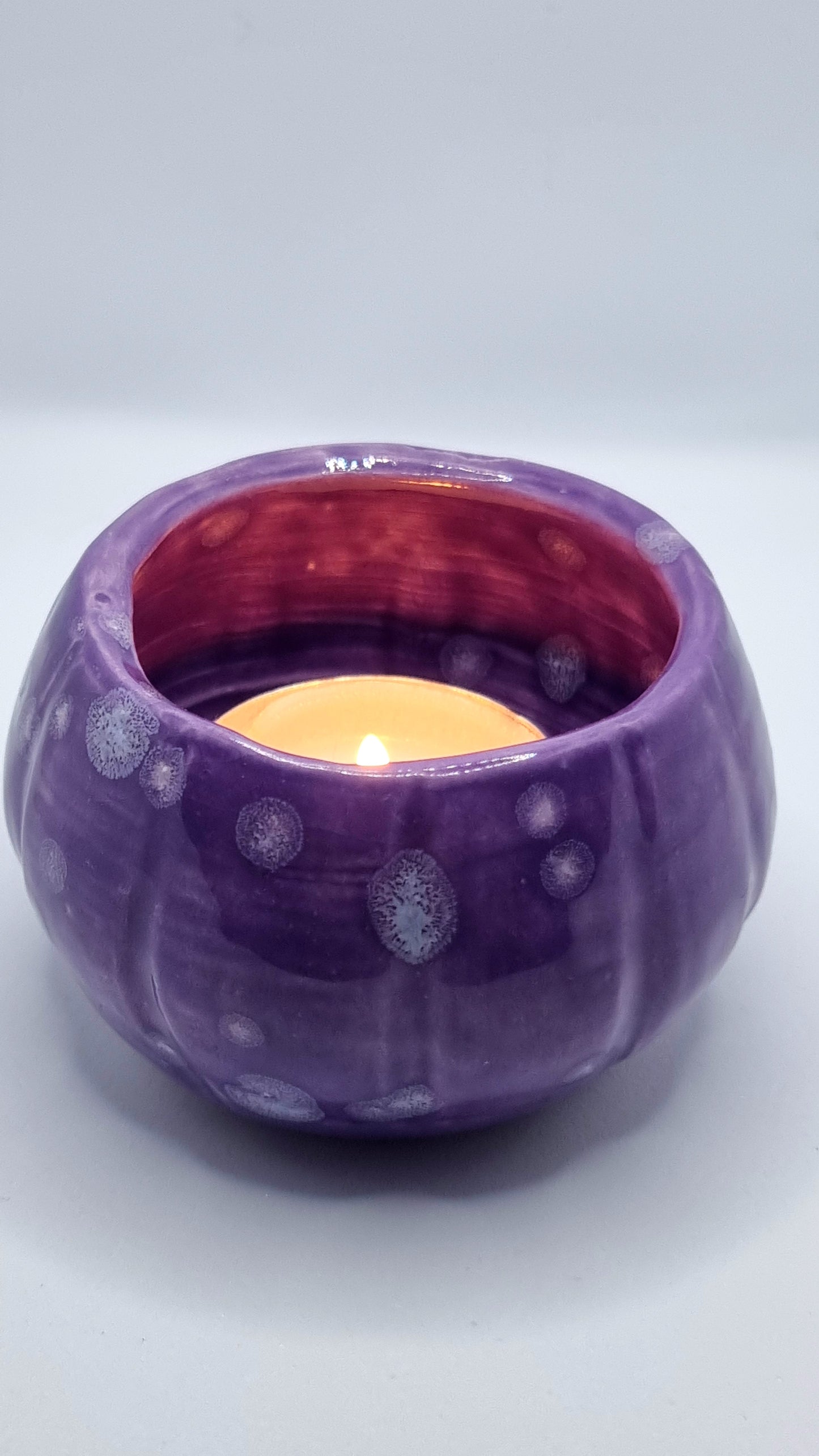 A purple, pumpkin-shaped tealight holder with a lit tealight inside on a neutral background 